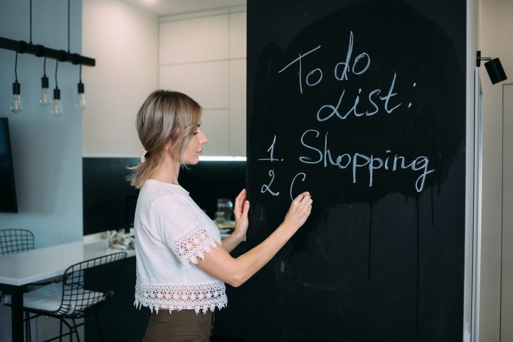 A blonde woman stands with her back and writes in chalk on a black board in the kitchen at home. To-do list in the kitchen. Mom is home alone.