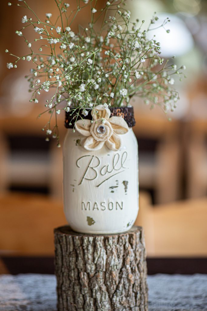 White mason jar centerpiece with canvass flower and babies breath inside on a wood stump