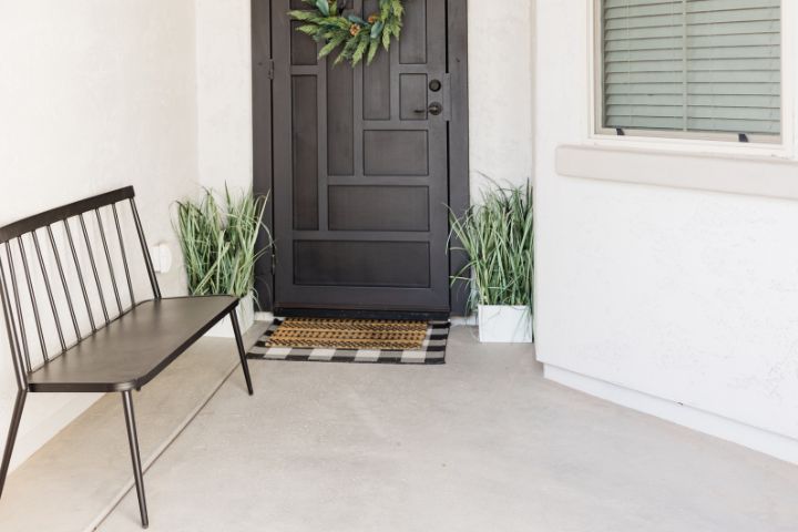 front porch at a beach home with two plants on either side of the door, a brown bench and a black door with decorative storm door.