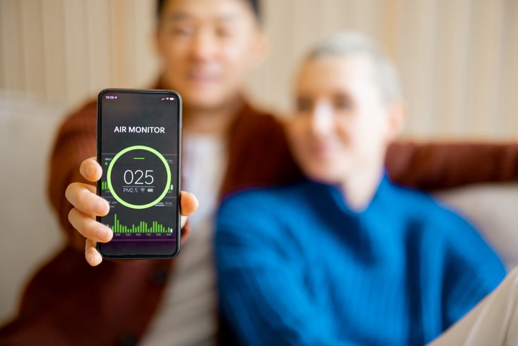 Asian man sitting on a sofa with his caucasian wife and showing a smartphone with a home air condition app. 