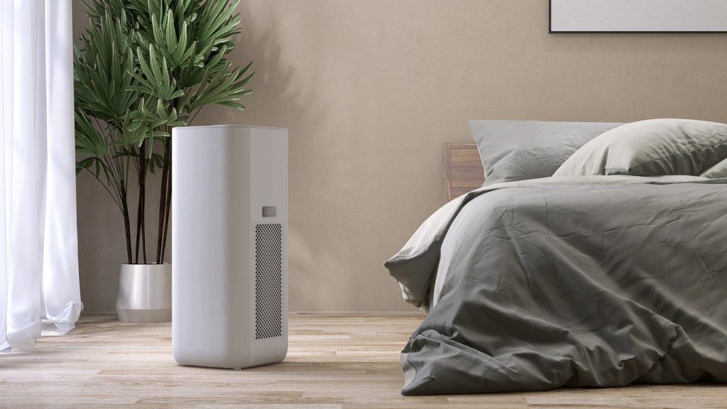 White modern design air purifier, dehumidifier in beige brown wall bedroom, gray cover sheet bed, tropical palm tree in sunlight on a wood floor 